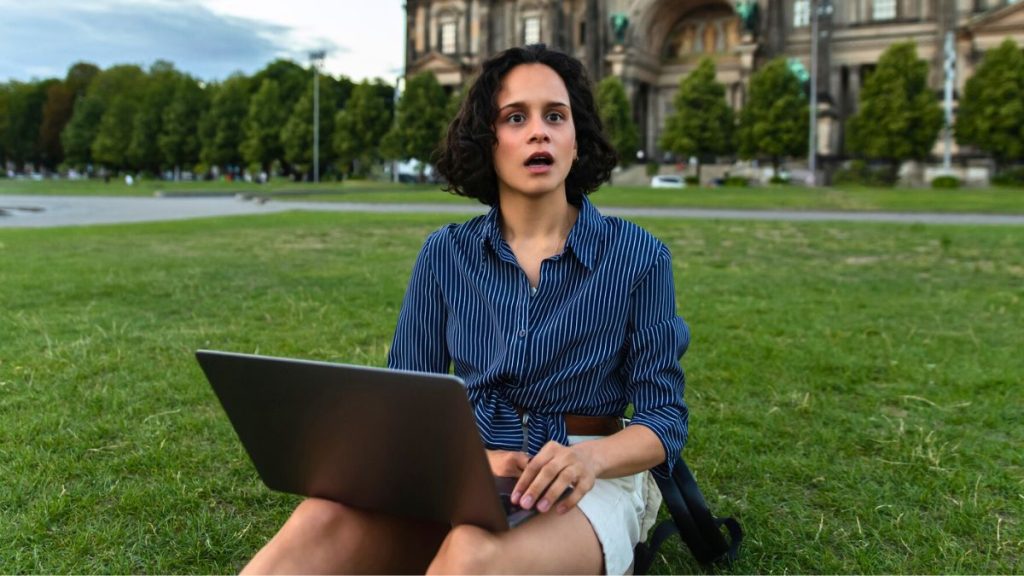 Shocked Young Woman Holding Laptop and Sitting on Grass MSN