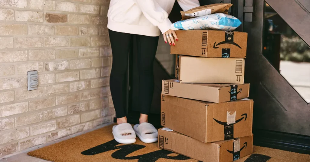 Things We Bought Today on Amazon That You Might Need woman standing next to a stack of amazon boxes on porch