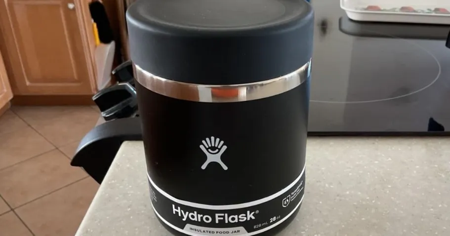 50% Off Hydro Flask Insulated Food Jars on Amazon A black Hydro Flask insulated food jar sits on a kitchen countertop near a stove. The jar has a silver accent and a logo.