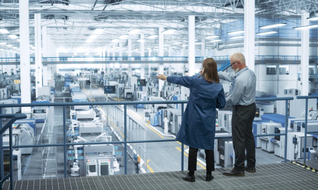 Experienced Male and Female Engineers Standing on a Platform with Their Back to Camera, Using Laptop Computer and Discussing Production at a Modern AI Automated Electronics Manufacture