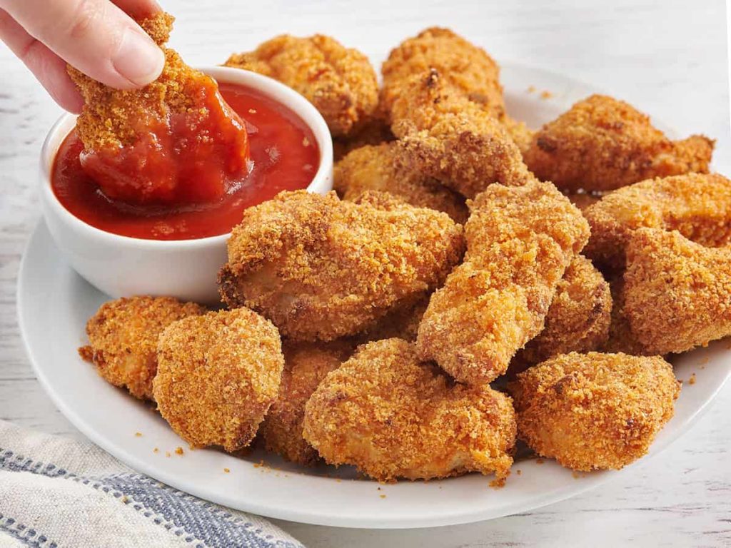 Side view of a plate of homemade chicken nuggets with a hand dipping a nugget into a pot of ketchup.