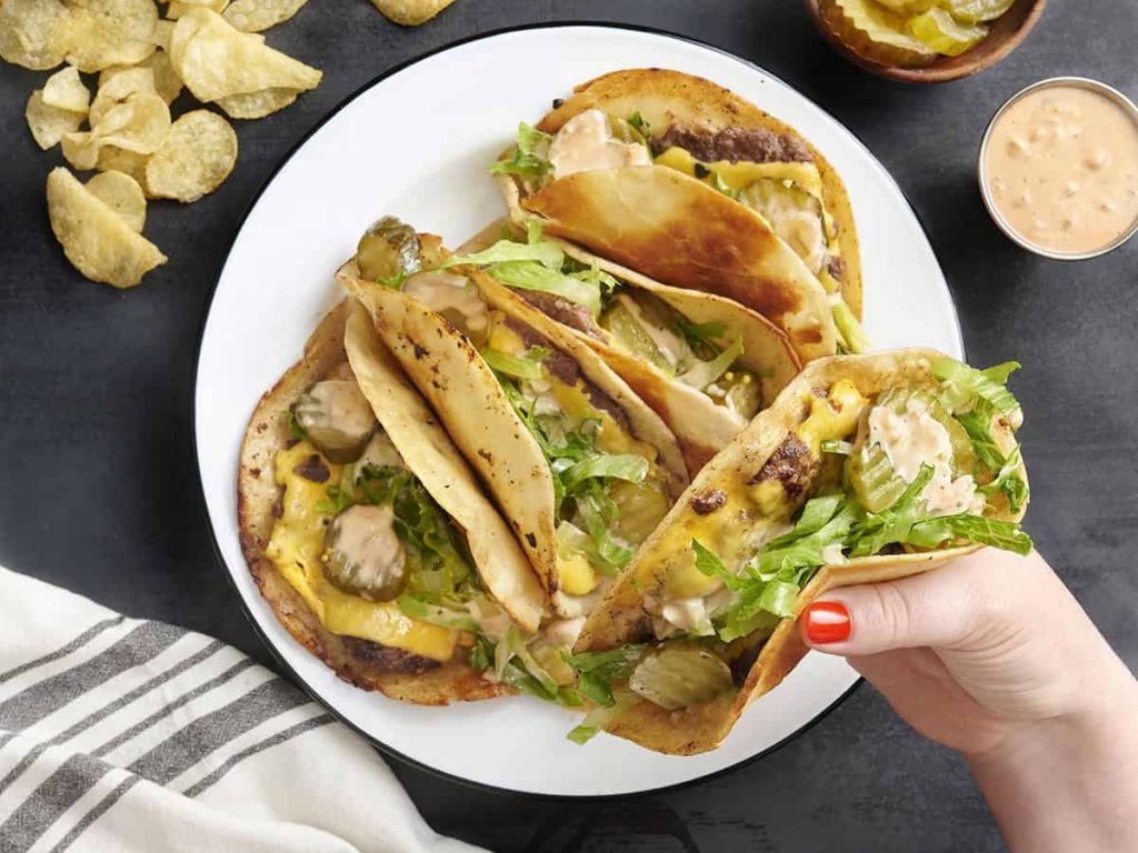 Overhead view of a plate of homemade smash burger tacos with a hand holding one.
