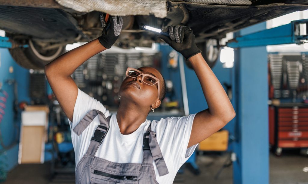 Mechanic Business Insurance: Companies, Costs and Coverage A young African American female mechanic is standing under a car and examining the damage on the broken parts with a serious look on her face.